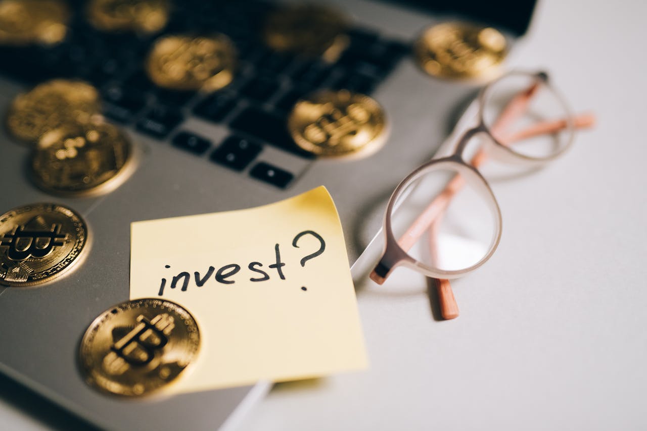 about-us-01 A close-up of bitcoin coins on a laptop with eyeglasses and a sticky note reading 'invest?'